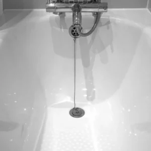 Black and white image of a bathtub with a central drain and a metal chain attached to it. A shower hose and faucet are mounted above the tub.