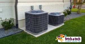 Two outdoor air conditioning units are placed on concrete slabs beside a house. The grass and small shrubs surround the area. A "Fetch-a-Tech" logo is visible in the bottom right corner.