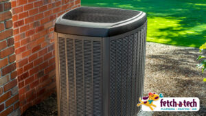 Outdoor air conditioning unit next to a brick wall in a garden, with a "fetch-a-tech" plumbing and heating logo in the bottom right corner.
