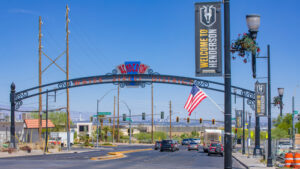 The entrance to Water Street District in Henderson boasts a steel arch adorned with an American flag and a welcome banner. As cars travel beneath the clear blue sky, nearby businesses offer top-notch HVAC and plumbing services to the community.