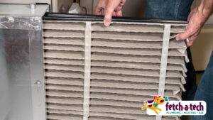 Person inserting an HVAC air filter into a unit, with a "fetch-a-tech" logo at the bottom right.